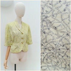 May include: A vintage, pale yellow blazer with a floral pattern and three buttons. The blazer has short sleeves and a notched collar. The fabric has a subtle texture, and the overall design is classic and elegant.