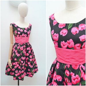 May include: A sleeveless black dress with a pink tulip floral pattern. The dress features a wide, solid pink waistband and a scoop neckline. The dress is displayed on a mannequin.