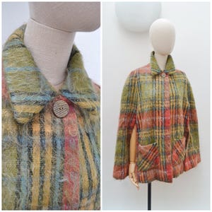 May include: A fuzzy, plaid cape featuring a blend of olive green, orange, yellow, and blue tones. This garment includes a pointed collar, a single brown button closure at the neck, and two front patch pockets. The fabric has a textured, mohair-like finish.