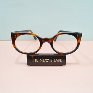 May include: Tortoiseshell pattern eyeglasses with star details on the upper corners. The glasses are displayed on a stand with the text "THE NEW SHAPE". The frames have a rounded, cat-eye shape.