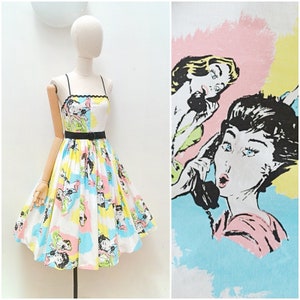 May include: A retro sundress with a comic book design in pastel shades. The dress has a white base with illustrations in black, yellow, pink, and blue. It features a black belt and thin straps. The dress is displayed on a mannequin.