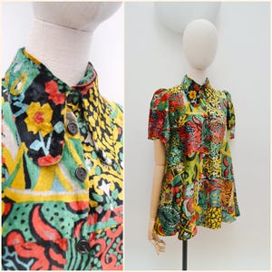 May include: A patterned short-sleeved blouse with a high collar and button-down front. The design incorporates red, yellow, green, and black elements. The blouse is displayed on a mannequin. The garment's design is eye-catching.
