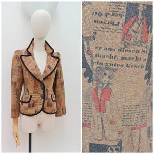 May include: A tan blazer featuring a repeating print of German text and illustrated figures in red and black. The jacket has a notched lapel, two front buttons, and black piping trim along the collar, lapels, pockets, and front edges.