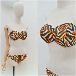 May include: A two-piece bikini set featuring a strapless bandeau top and matching bikini bottoms. The swimwear displays a bold, abstract graphic pattern in vibrant shades of orange, yellow, black, and white, creating a striking visual contrast.