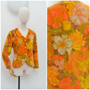 May include: A long-sleeve blazer featuring a vibrant floral pattern in shades of orange, yellow, and brown. The garment has a V-neckline, three matching orange buttons down the front, and a soft, textured finish on the fabric surface.