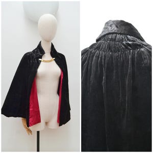 May include: A black velvet cape with a red satin lining, displayed on a mannequin. The cape features a high collar and is fastened with a gold-toned clasp. The fabric has a luxurious sheen, and the cape drapes elegantly.