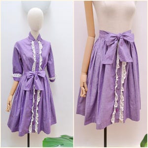 May include: A lavender purple dress and a matching skirt, both featuring a button-down front, a vertical white lace trim, and a fabric waist tie. The dress has three-quarter length sleeves with lace cuffs, while the skirt has a high-waisted design.