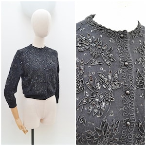May include: A black beaded cardigan with a round neckline and long sleeves. The vintage sweater features intricate floral and leaf patterns with silver sequins and beads. The cardigan is displayed on a mannequin.