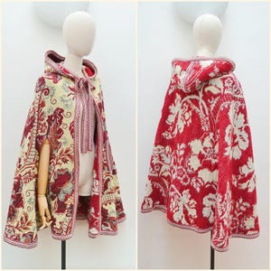 May include: A hooded cape with a floral pattern in red, cream, and burgundy. The cape features a decorative trim and a tie closure at the neck. The design is displayed on a mannequin, showcasing the garment's shape and details.