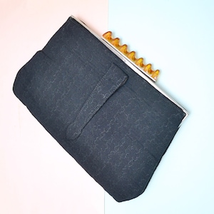 May include: A black rectangular clutch bag featuring a textured fabric surface with a subtle repeating pattern. The bag has a silver-toned metal frame and a decorative amber-coloured resin clasp with a scalloped, wave-like edge design.