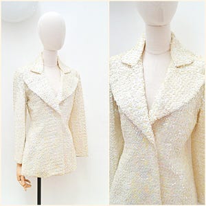 May include: A cream-coloured sequined blazer with a classic lapel collar and long sleeves. The jacket is displayed on a mannequin, highlighting its tailored fit and textured surface. The sequins add a subtle shimmer to the garment.