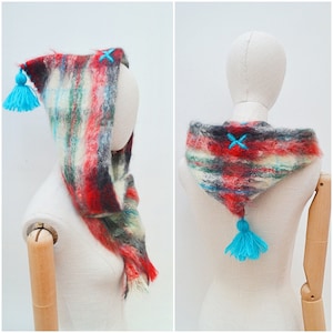 May include: A colorful, handmade hood scarf with a tassel. The scarf features a blend of red, black, white, and teal fibers. It has a hood with a teal tassel and teal cross-stitch details.
