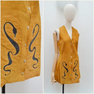 May include: Mustard yellow suede vest dress with a V-neck and button closure. The dress features black snake-like designs on the front. The dress is displayed on a mannequin.