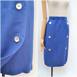 May include: A cobalt blue skirt with a wrap design and a row of six decorative buttons. The skirt is made of a solid-colored fabric and has a tailored fit. The buttons are round and have a pearlescent finish.