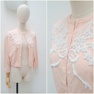May include: A pale peach-coloured cardigan featuring a round neckline, three-quarter length sleeves, and a front button closure. The garment is embellished with intricate white beadwork arranged in floral and ribbon-like patterns across the chest.