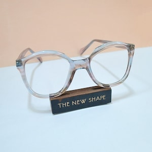 May include: A pair of eyeglasses with a light pink and blue marbled frame, resting on a small wooden block. The block has the text "THE NEW SHAPE" in white. The glasses have a rounded, cat-eye shape.