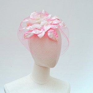 May include: A pink and white floral headpiece with a fine pink netting veil. The headpiece features fabric leaves and a central flower, in shades of pink and white. The veil adds a touch of elegance to this decorative accessory.