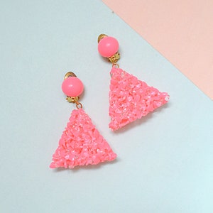 May include: A pair of clip-on earrings featuring a round, smooth pink bead at the top connected to a gold-tone metal finding. Below, a triangular pendant is covered in small, textured, bright pink crushed stone-like fragments.