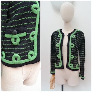 May include: A black long-sleeve cardigan featuring horizontal green stripes, a round neckline, and green braided trim along the front opening and pockets. The garment includes three black circular buttons and green cuffs on the sleeves.