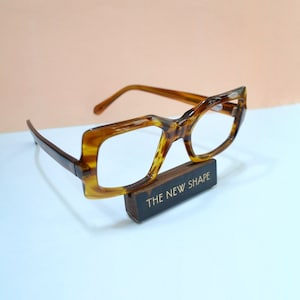 May include: Brown tortoiseshell eyeglasses with a unique, angular frame design. The glasses rest on a small wooden block with the text "THE NEW SHAPE" in white. The frames have a retro, vintage aesthetic.