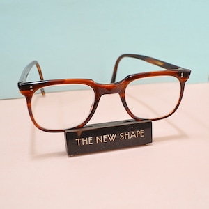 May include: Brown tortoiseshell eyeglasses with a rectangular frame, resting on a black stand. The stand has the text "THE NEW SHAPE" in gold lettering. The background is a light blue and pink.