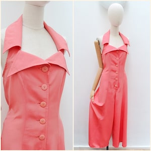 May include: A coral-coloured sleeveless jumpsuit featuring a wide collar, a button-down front with six matching circular buttons, and wide-leg trousers. The garment is displayed on a mannequin, showcasing the structured bodice and collar design.