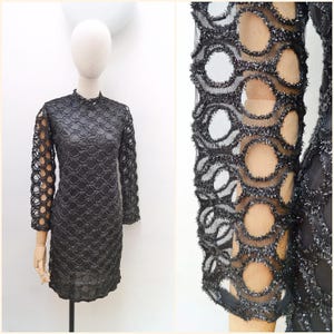 May include: A black, knee-length dress with a high neckline and long sleeves. The dress features a unique openwork design with circular cutouts and a shimmering, textured trim. The dress is displayed on a mannequin.