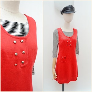 May include: A red sleeveless dress with silver buttons and two small pockets. It is worn over a black and white striped long-sleeved top. The dress is displayed on a mannequin. The mannequin is wearing a black cap.
