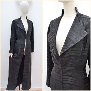 May include: A long, black coat with a textured pattern, displayed on a mannequin. The coat features a notched collar and a long, open front. The fabric has a subtle, repeating design.