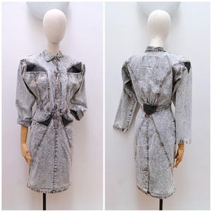 May include: A acid-wash denim dress featuring long sleeves, a button-down front, and black triangular fabric inserts at the shoulders and waist. The garment has a structured collar, defined waistline, and a knee-length skirt with visible seams.
