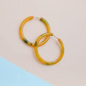 May include: A pair of hoop earrings. The earrings are a translucent yellow with green and black speckles. The earrings have a silver post and are set against a pink and blue background.