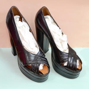 May include: A pair of black platform heels with a peep-toe design. The shoes have thick, block heels and a criss-cross strap across the top. The shoes are filled with white paper and sit on a light blue surface.