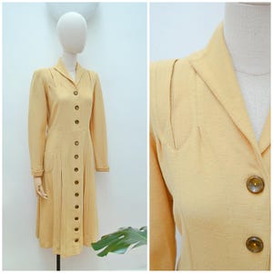 Könnte beinhalten: Ein hellgelbes Vintage-Kleid mit Knopfleiste, langen Ärmeln und einem Kragen. Das Kleid hat eine taillierte Passform mit Falten am Rock und dekorativen Schulterdetails. Das Kleid wird auf einer Schaufensterpuppe präsentiert.