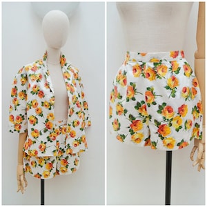 May include: A two-piece set consisting of a short-sleeved button-down shirt and matching shorts, both featuring a white base fabric with a repeating pattern of yellow and orange roses with green leaves, displayed on a mannequin.