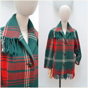 May include: A long-sleeved jacket featuring a green and red plaid pattern with white accent lines. The jacket has a notched collar with fringe detailing, three dark brown buttons down the front, and a fringed hemline at the bottom edge.