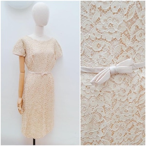 May include: A cream-colored lace dress featuring short sleeves, a round neckline, and a slim silhouette. The dress is accented with a thin, matching cream-colored ribbon tied into a small bow at the waist, layered over a solid cream-colored lining.