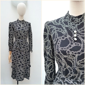 May include: A vintage black dress with a repeating chain-link pattern in a lighter color. The dress features long sleeves, a high neckline with three white buttons, and a matching belt. The dress is displayed on a mannequin.
