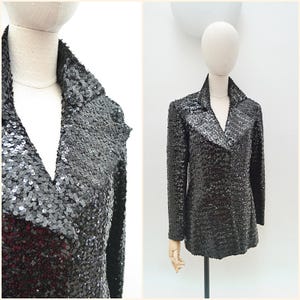 May include: A black sequin blazer with a notched collar and long sleeves. The jacket is displayed on a mannequin, showcasing its shimmering texture and tailored fit. The sequins create a dazzling effect.