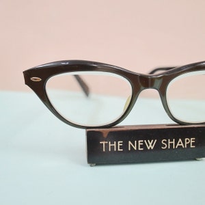 May include: Dark brown cat-eye eyeglasses with clear lenses. The glasses have a gold accent on the outer corners. The glasses are displayed on a wooden stand with the text "THE NEW SHAPE" in gold lettering. The background is a light pink and blue.