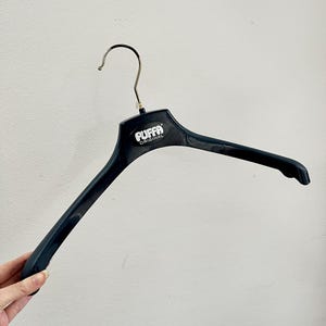 May include: A black plastic clothes hanger featuring a silver metal hook at the top. The center of the hanger displays the white text "PUFFA ORIGINAL" printed on a raised oval section. The hanger has wide, contoured arms for garment support.