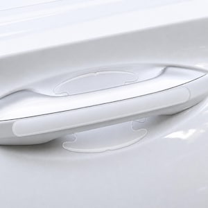 May include: A clear, adhesive protective film applied to a white car door handle and the surrounding recessed area. The transparent material is shaped to fit the contours of the handle and the cup, providing a layer of protection against scratches.