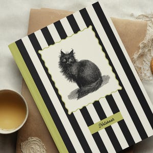 May include: Notebook with a black and white striped cover, featuring a framed illustration of a black cat. The spine is lime green, and a small label reads "Notebook". A cup of tea and a cookie are also present.