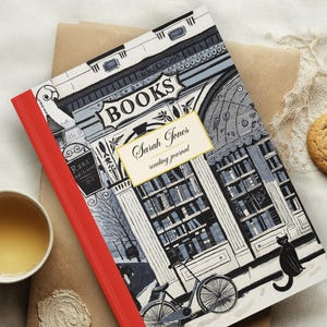 May include: A hardcover reading journal with a red spine and a detailed illustration of a bookstore facade. The cover reads "BOOKS" and "Sarah Jones reading journal." A cup of tea and a cookie are also present.