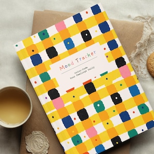 Puede incluir: Un diario de seguimiento del estado de ánimo con un patrón de cuadros amarillo, naranja, azul, negro y verde. La portada dice "Mood Tracker" y "Color-code Your Feelings Daily". Una taza de té y una galleta están al lado del diario.
