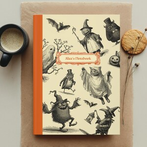 May include: A notebook with an orange spine and a cream-coloured cover featuring whimsical Halloween illustrations. The cover includes the text "Alice's Notebook" in a decorative frame. A cup of coffee and a biscuit are also in the image.