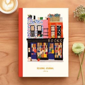 May include: A Reading Journal with a colorful illustration of a bookstore facade. The cover features a red spine and the words "BOOKS" above the bookstore illustration. A cup of coffee and a flower are next to the journal on a wooden surface.
