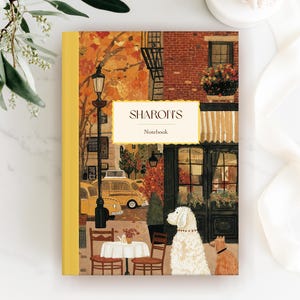 Puede incluir: Un cuaderno con lomo amarillo y una portada con una escena callejera ilustrada. La escena incluye un coche amarillo, un café con mesas y sillas y dos perros. El texto "SHARON'S Notebook" se muestra en la portada.