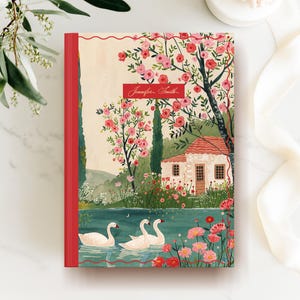 Op de afbeelding: Een hardcover journal met een rode rug en een landschapsillustratie. De omslag toont een serene landschap met een meer, zwanen, een klein huis en bloeiende bomen. De naam "Jennifer Smith" staat op een rode banner.