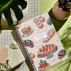 May include: Spiral-bound notebook with a cream-colored cover featuring colorful illustrations of pastries and a teacup. The notebook is labeled "Jennifer's notebook" in a pink rectangle. A decorative notebook for writing and journaling.