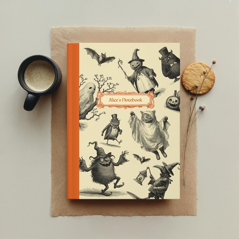 May include: A notebook with an orange spine and a cream-coloured cover featuring whimsical Halloween illustrations. The cover includes the text "Alice's Notebook" in a decorative frame. A cup of coffee and a biscuit are also in the image.